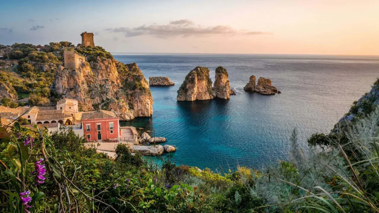 Un borgo con torri antiche e una casa rossa si affaccia su un mare bello Italia, con faraglioni che emergono dalle acque cristalline.