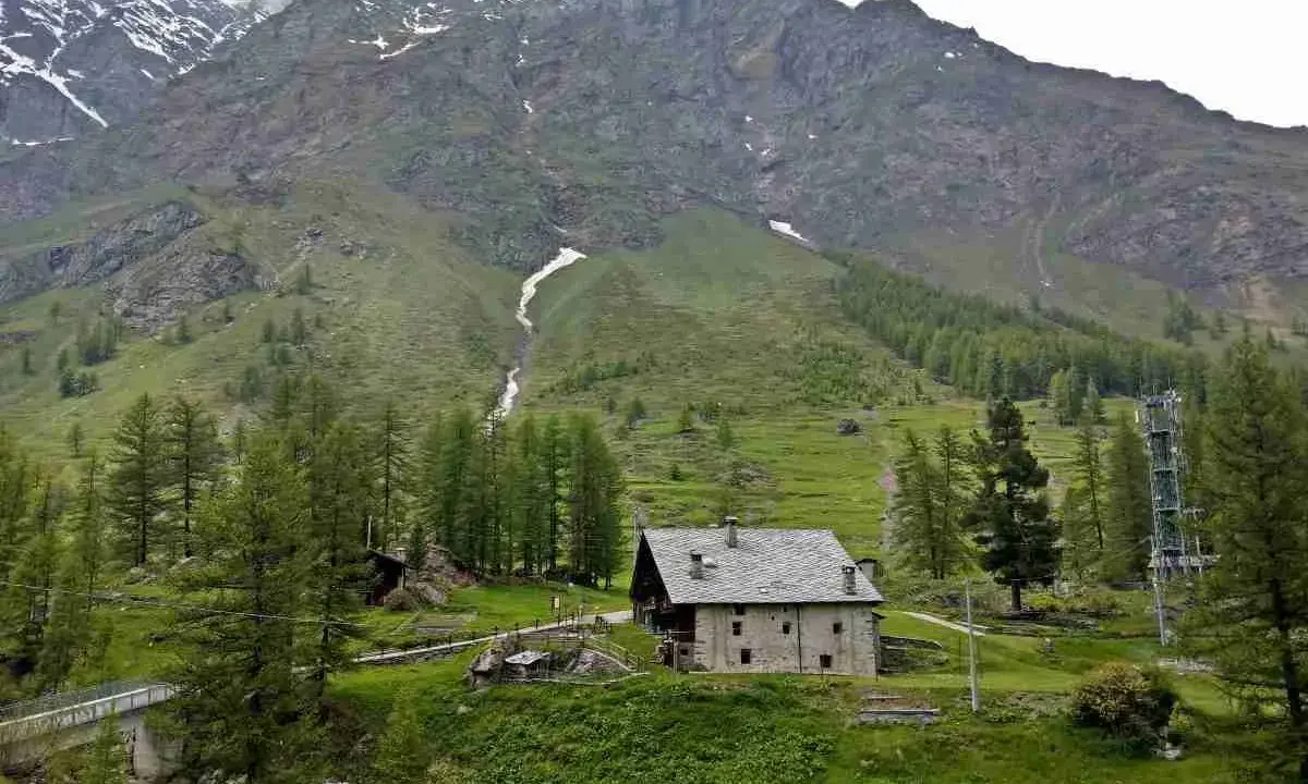 Un casolare in pietra nel **Val di Rhemes**, circondato da verdi pendii e imponenti montagne innevate.