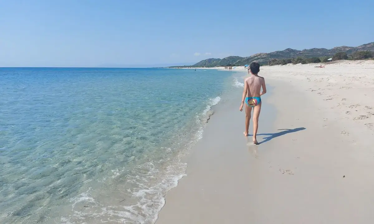 Un bambino cammina su una spiaggia di sabbia bianca, verso le spiagge più belle della Sardegna est. Acqua cristallina e cielo azzurro.