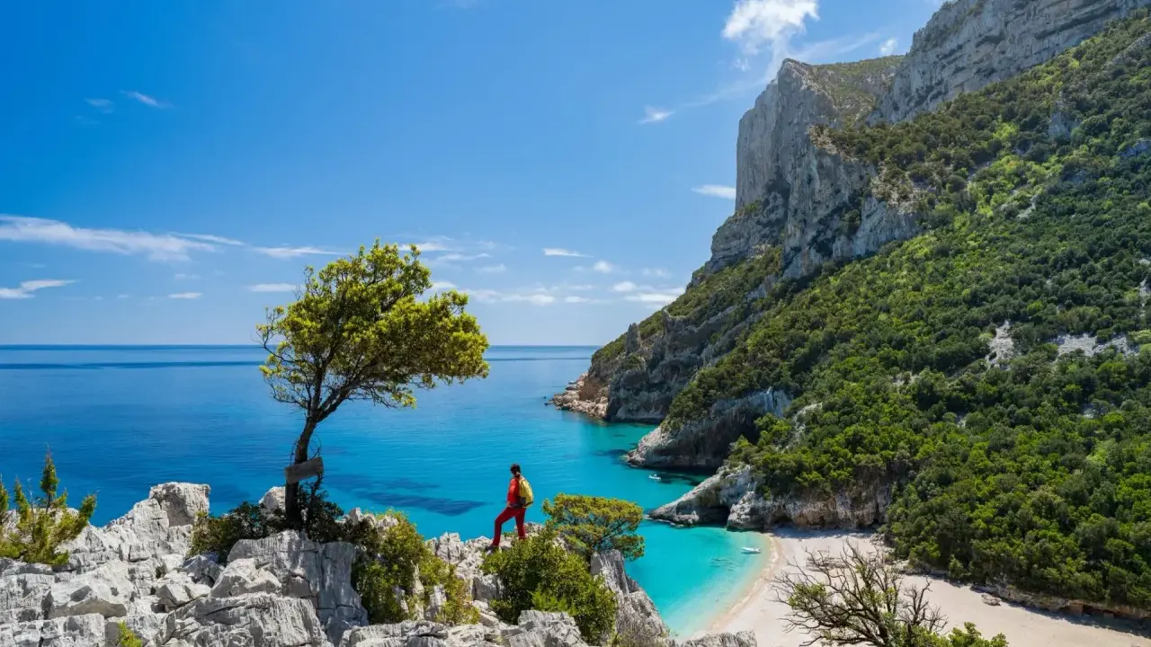 Escursionista ammira una baia cristallina con una spiaggia bianca, incastonata tra imponenti scogliere verdi. Un paesaggio mozzafiato che ricorda la bellezza della montagna centro Italia.