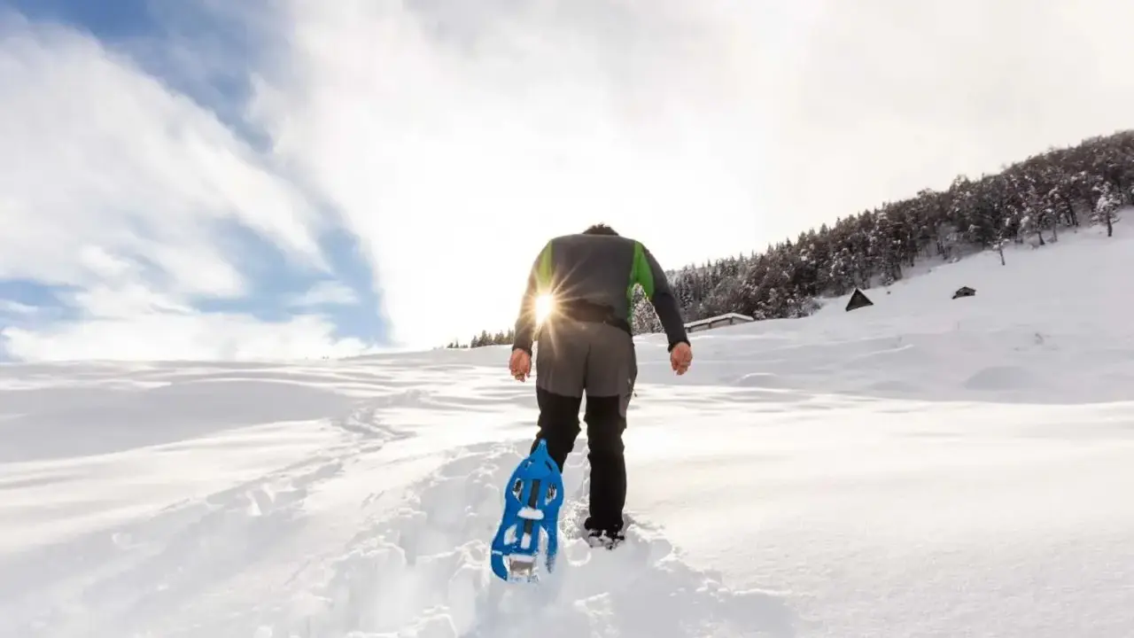 Uomo con ciaspole cammina su sentiero innevato, ideale per chi cerca dove ciaspolare in Piemonte. Sole splendente tra le nuvole.
