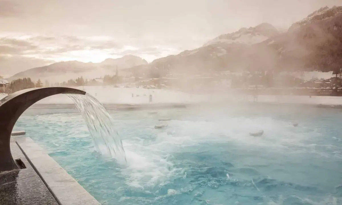 Una cascata d'acqua calda scende in una piscina all'aperto, con montagne innevate e un villaggio sullo sfondo. Un'oasi di relax a Pozza di Fassa QC Terme.