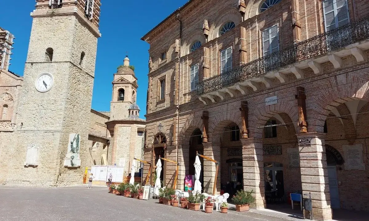 Piazza a San Ginesio, Marche, con il campanile in pietra e l'edificio in mattoni rossi con portici.
