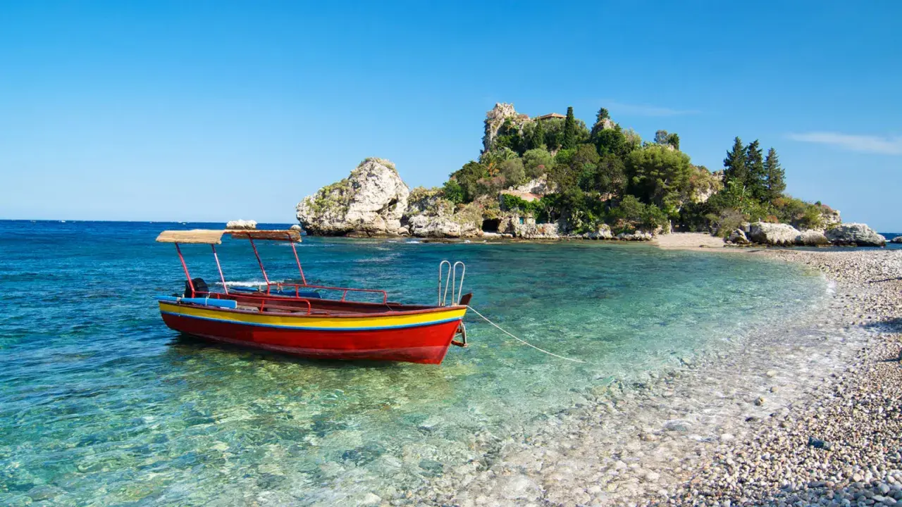 Barca rossa e gialla in acque cristalline, con isolotto verdeggiante sullo sfondo. Un angolo di paradiso che ricorda la bellezza di Sicilia e Sardegna.