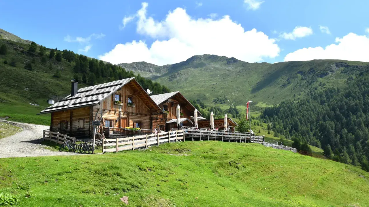 Malga Alto Adige, un rifugio in legno con tavoli all'aperto, circondato da verdi pendii e montagne boscose sotto un cielo azzurro con nuvole bianche.