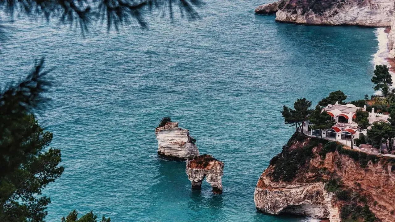 Scogliere mozzafiato e spiagge deserte Italia, con faraglioni che emergono dal mare turchese e una villa panoramica.