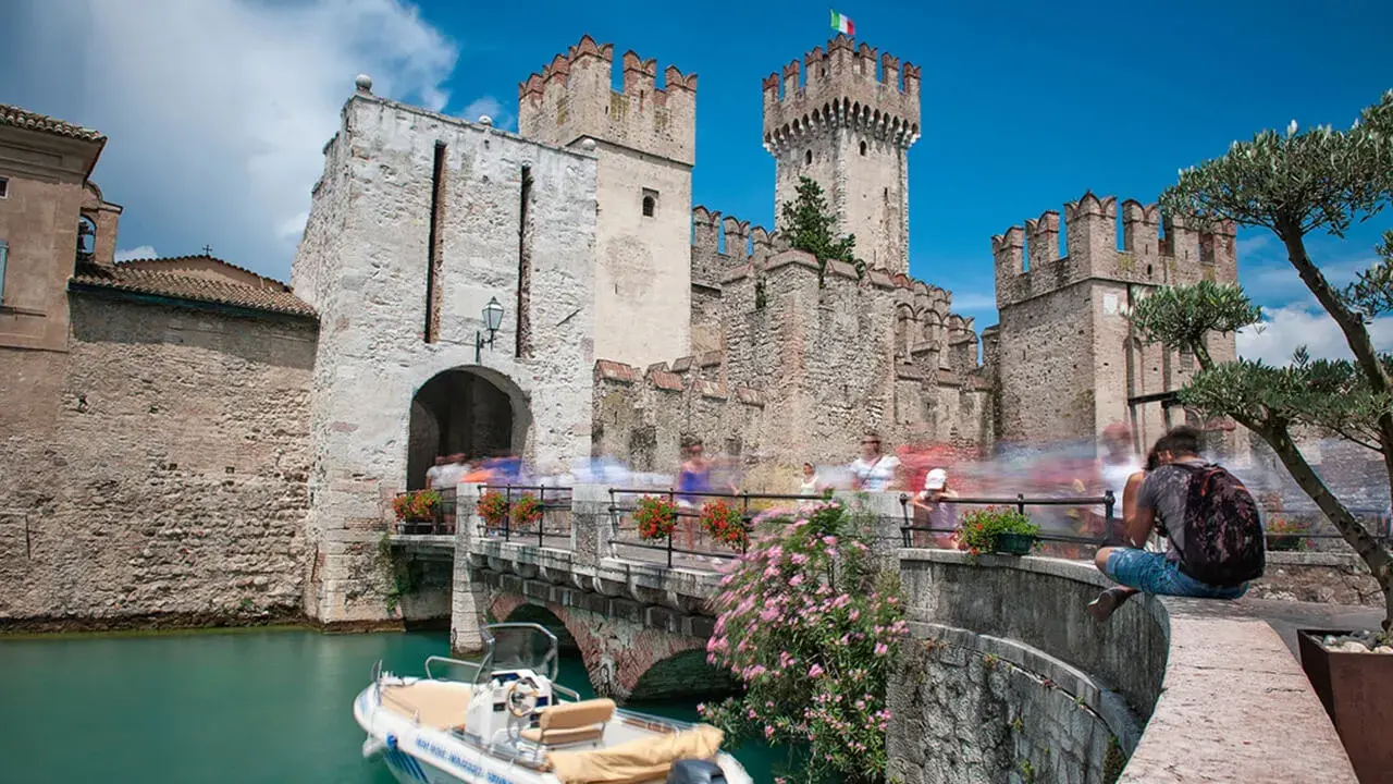 Castello Scaligero di Sirmione, un'ottima idea per cosa vedere a Sirmione in un giorno. Un ponte pedonale affollato porta all'ingresso del castello, con un motoscafo ormeggiato nell'acqua turchese.