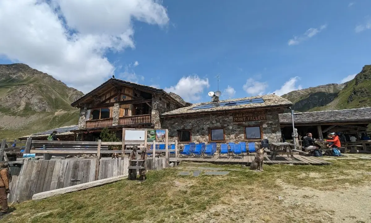 Rifugio Mont Fallère, uno dei rifugi Valle d'Aosta facili, accoglie escursionisti con le sue strutture in pietra e legno, circondato da verdi pendii e montagne.