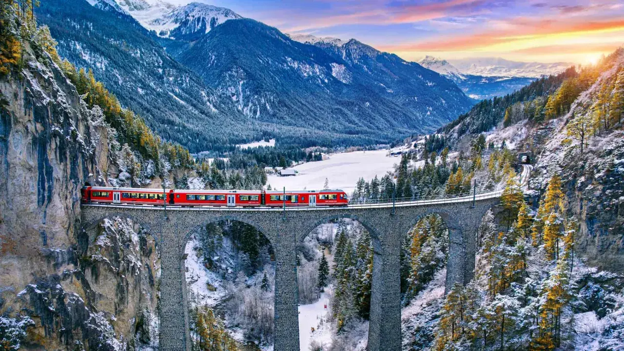 Il trenino del Bernina attraversa un viadotto in pietra in un paesaggio alpino innevato, con montagne maestose e un cielo colorato al tramonto.