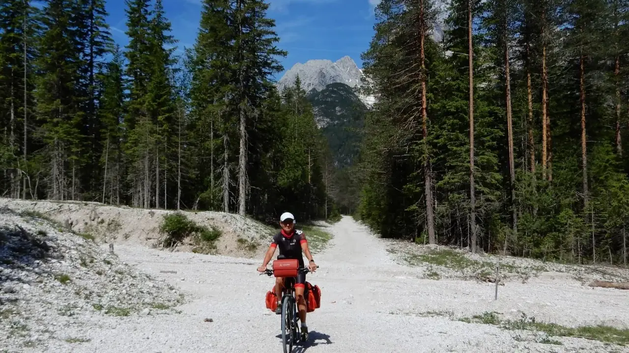 Ciclista su strada sterrata tra i pini, con le vette delle Dolomiti sullo sfondo. Un'avventura sui percorsi bici Dolomiti.