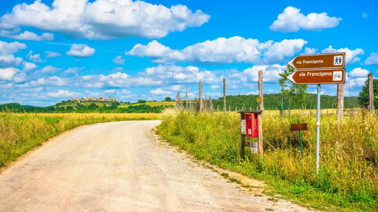 Segnaletica per la Via Francigena, un antico cammino. La strada sterrata conduce verso un borgo collinare sotto un cielo azzurro con nuvole bianche.