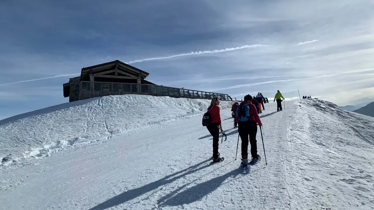 Gruppo di escursionisti con ciaspole su un sentiero innevato, con un rifugio in legno in cima a una collina. Posti belli in montagna Piemonte inverno.
