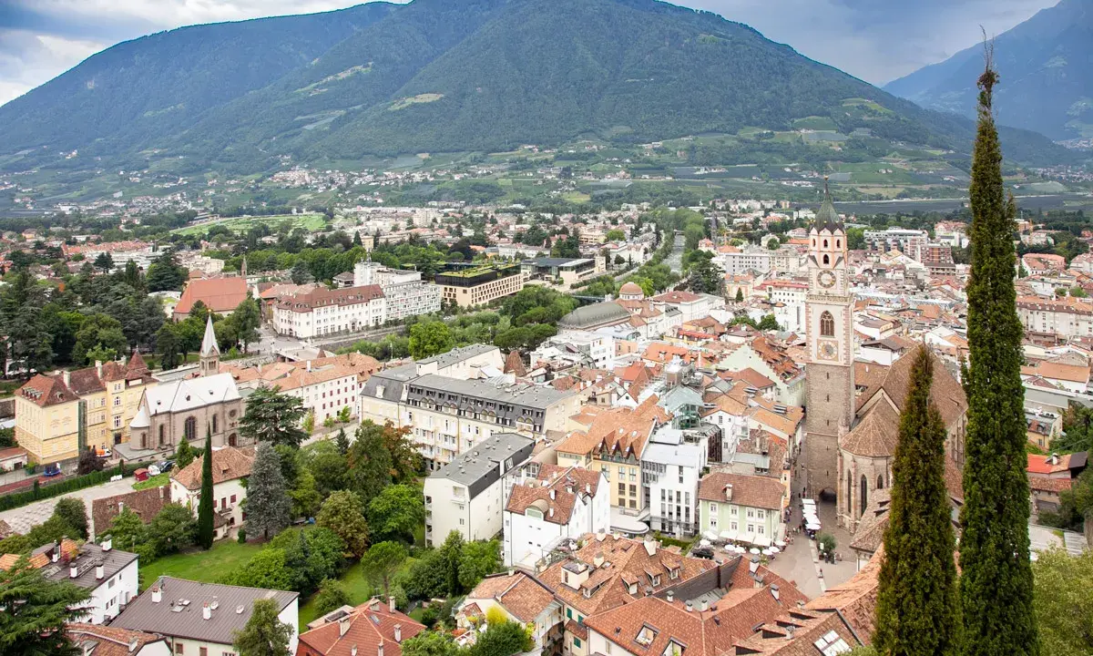 Vista aerea del centro storico di Merano, con montagne sullo sfondo e un campanile che svetta tra i tetti rossi.