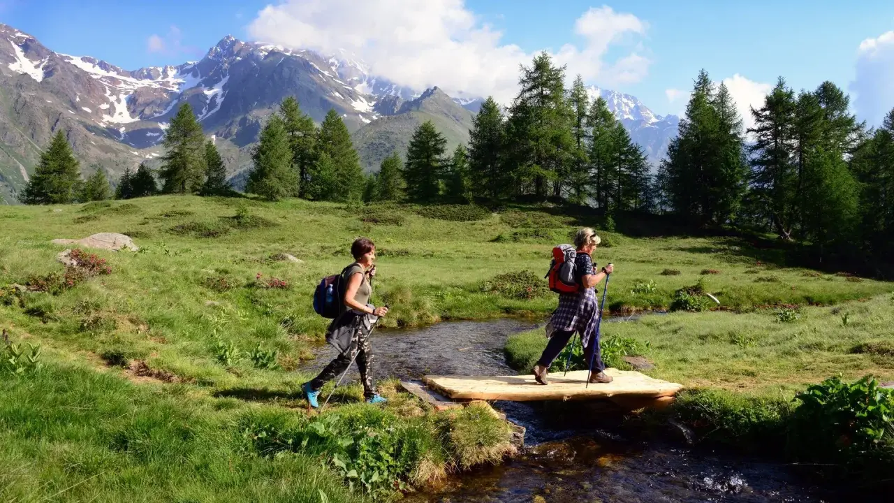Due escursioniste attraversano un ruscello su un ponte di legno, godendosi un'attività in montagna tra prati verdi e cime innevate.