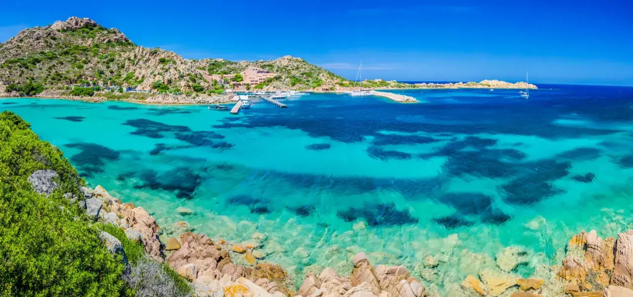 Acque cristalline e rocce granitiche ti aspettano per una visita alla Maddalena. Un paradiso azzurro con barche all'ancora.