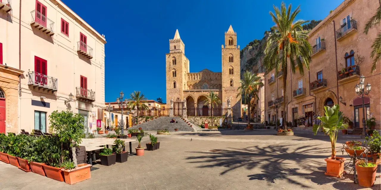 Piazza soleggiata con la cattedrale normanna sullo sfondo, palme e palazzi storici. Un luogo incantevole tra i borghi da visitare in Sicilia.