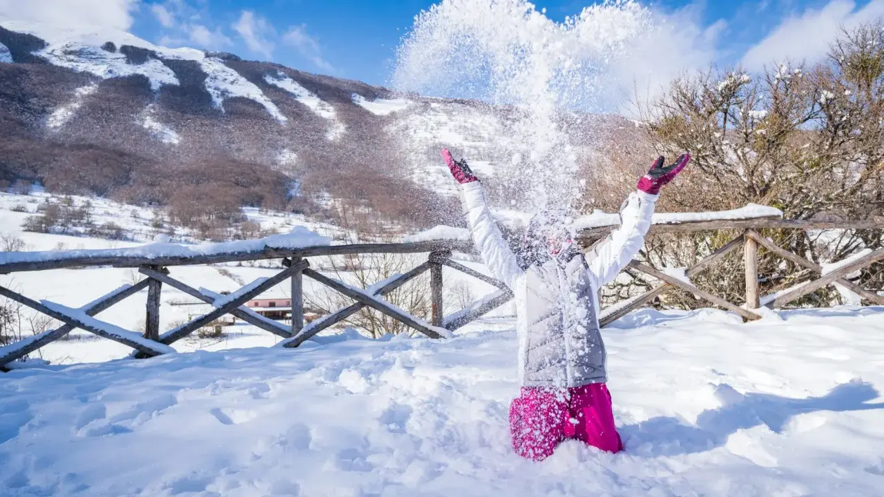 Bambina felice lancia neve in aria, con montagne innevate sullo sfondo. Perfetto per scoprire dove andare per la neve in Abruzzo.