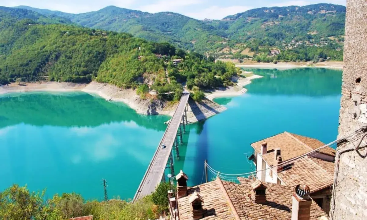 Panoramica mozzafiato del lago turchese vicino a Castel di Tora, con un ponte che attraversa le acque cristalline e montagne verdi sullo sfondo.