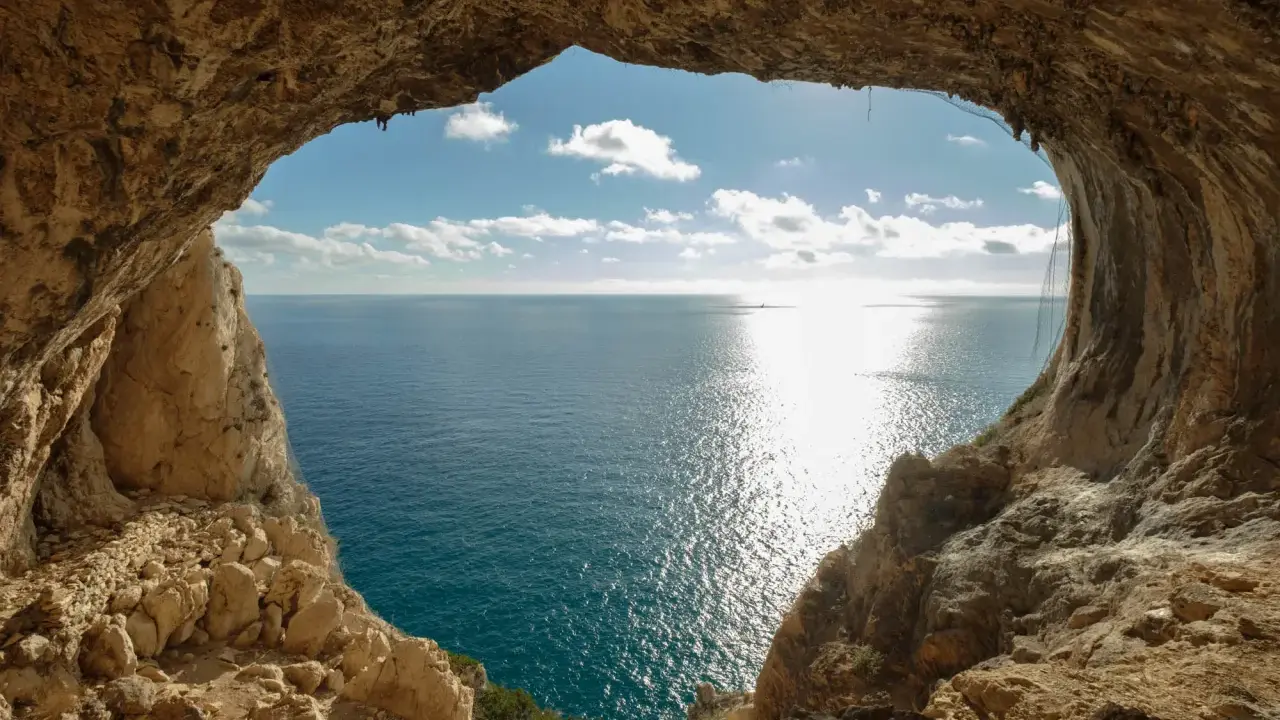 Vista dal mare da una grotta, con un sentiero del pellegrino che si intravede tra le rocce. Il sole brilla sull'acqua.