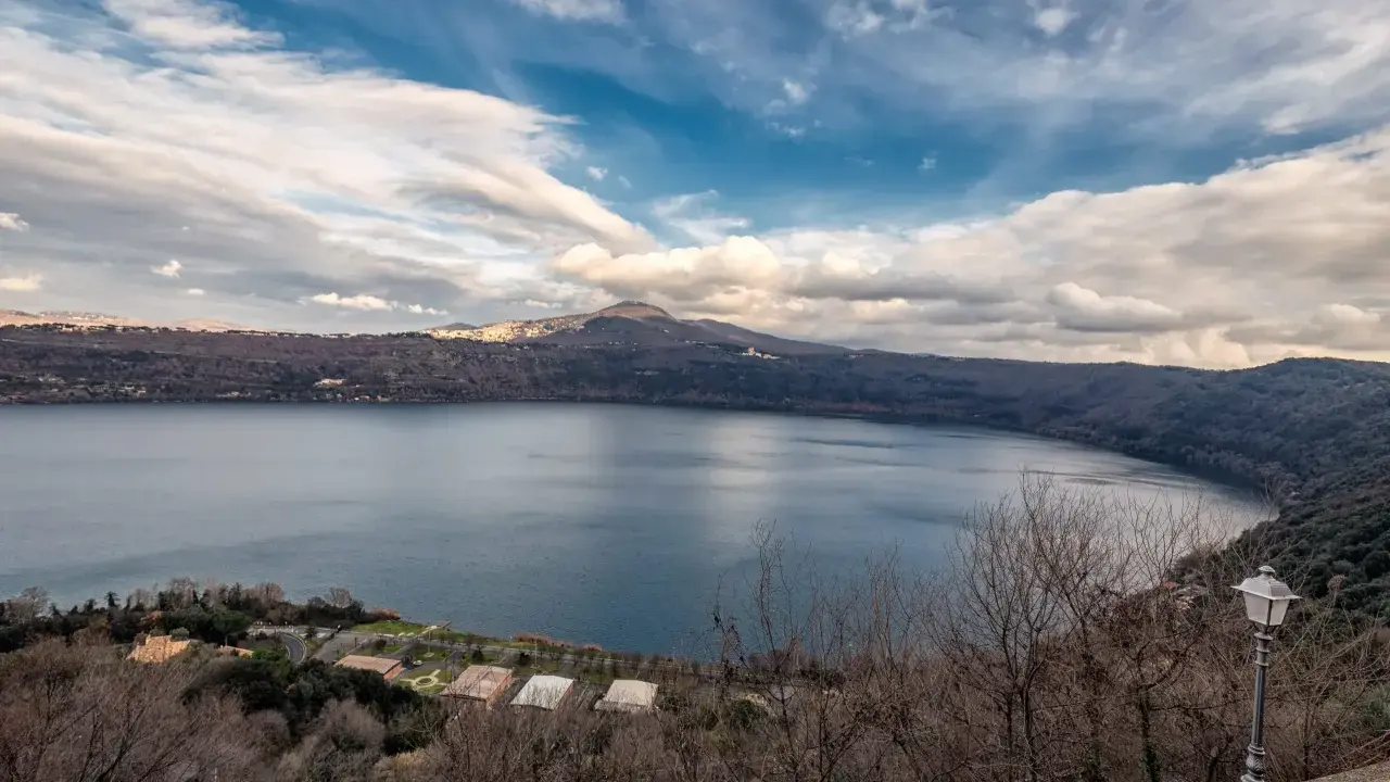 Vista panoramica del lago di Castel Gandolfo, Italia, con colline boscose e un cielo nuvoloso.