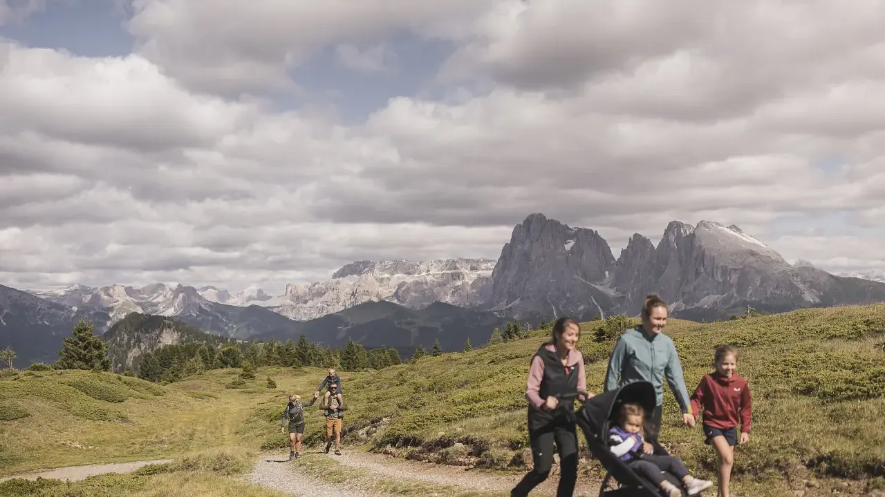 Famiglia in escursione sull'Alpe di Siusi, con bambini e passeggino, sullo sfondo le maestose vette dolomitiche.