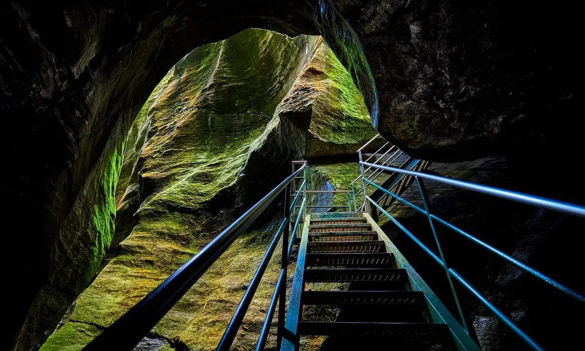 Una scala metallica sale tra le rocce muschiose degli orridi di Uriezzo, illuminata da una luce che filtra dall'alto.