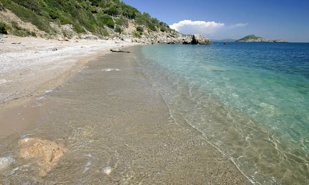Spiaggia di ciottoli e sabbia con acqua cristallina, vista verso il mare aperto e un'isola in lontananza, vicino al porto di Ercole.