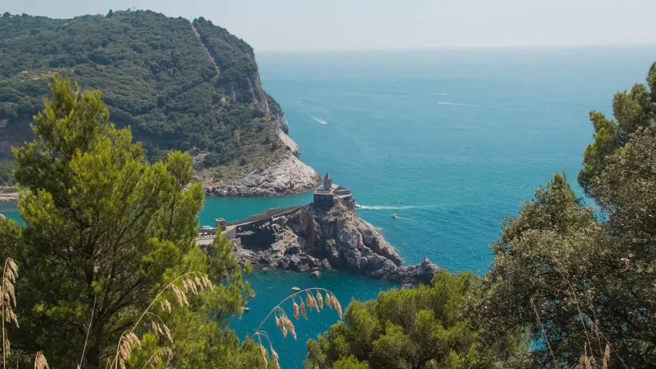 Vista panoramica del sentiero Riomaggiore Portovenere, con la chiesa di San Pietro su uno sperone roccioso e il mare azzurro.