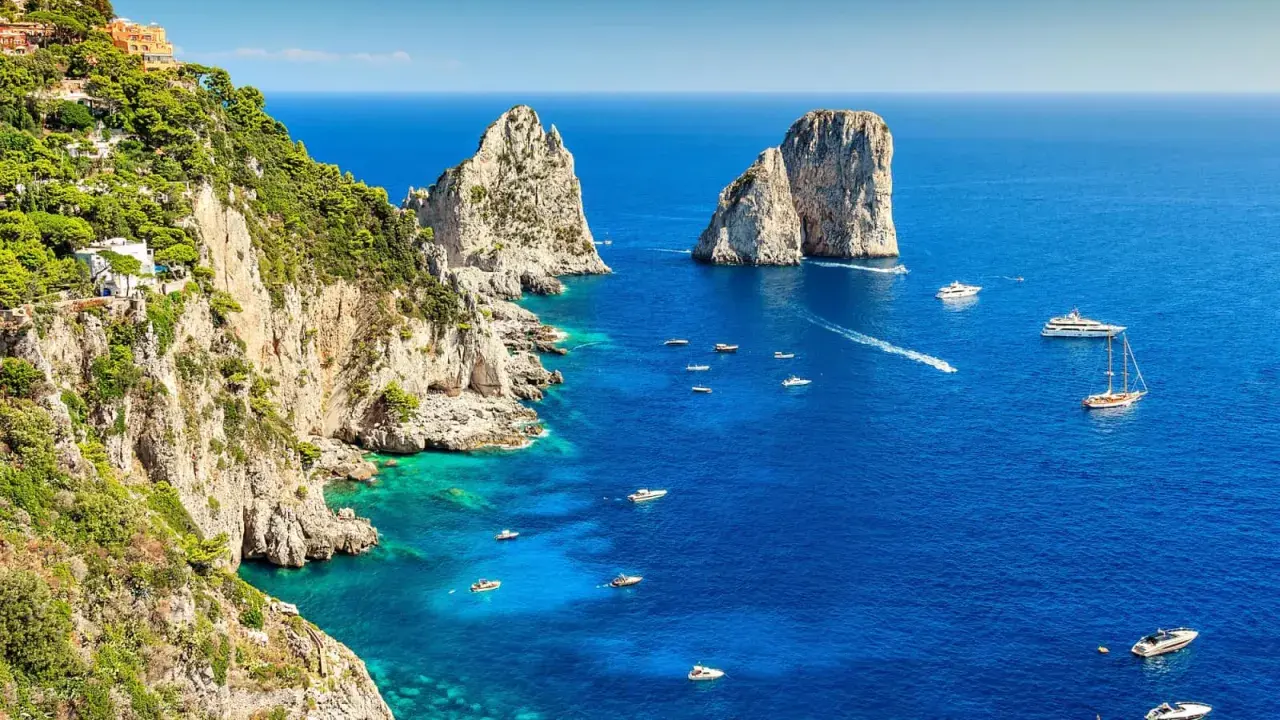 Scogliere verdi e rocciose si affacciano sul blu intenso del golfo di Napoli, punteggiato da barche e yacht. Faraglioni iconici emergono dal mare.
