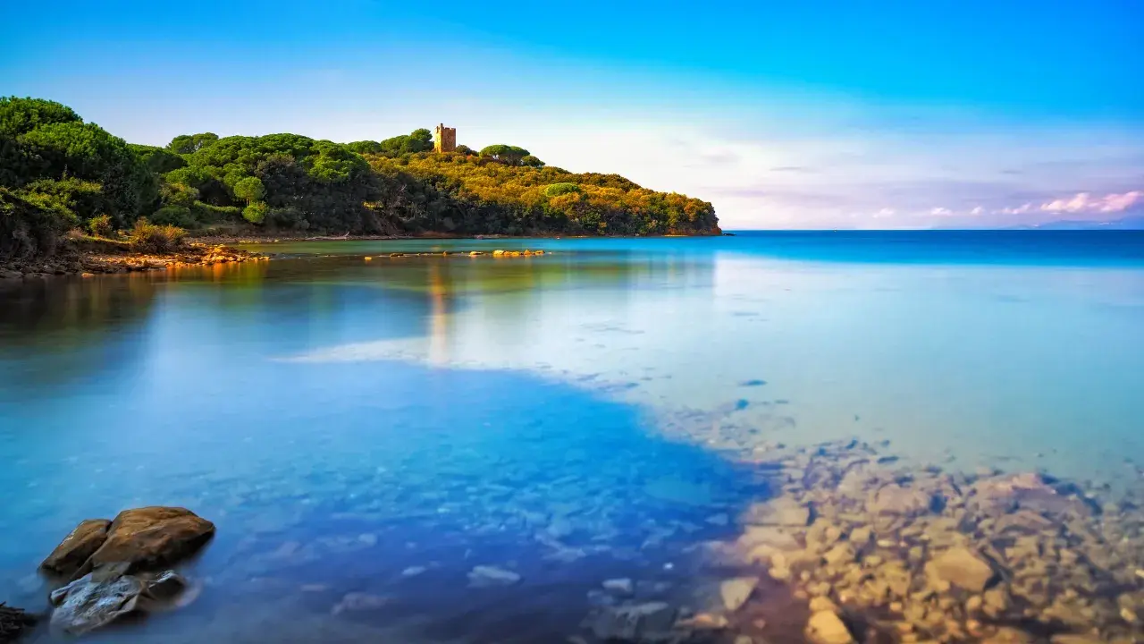 Torre medievale su una collina verdeggiante affacciata sul limpido **golfo di Follonica**. Acque calme e trasparenti riflettono il cielo azzurro.