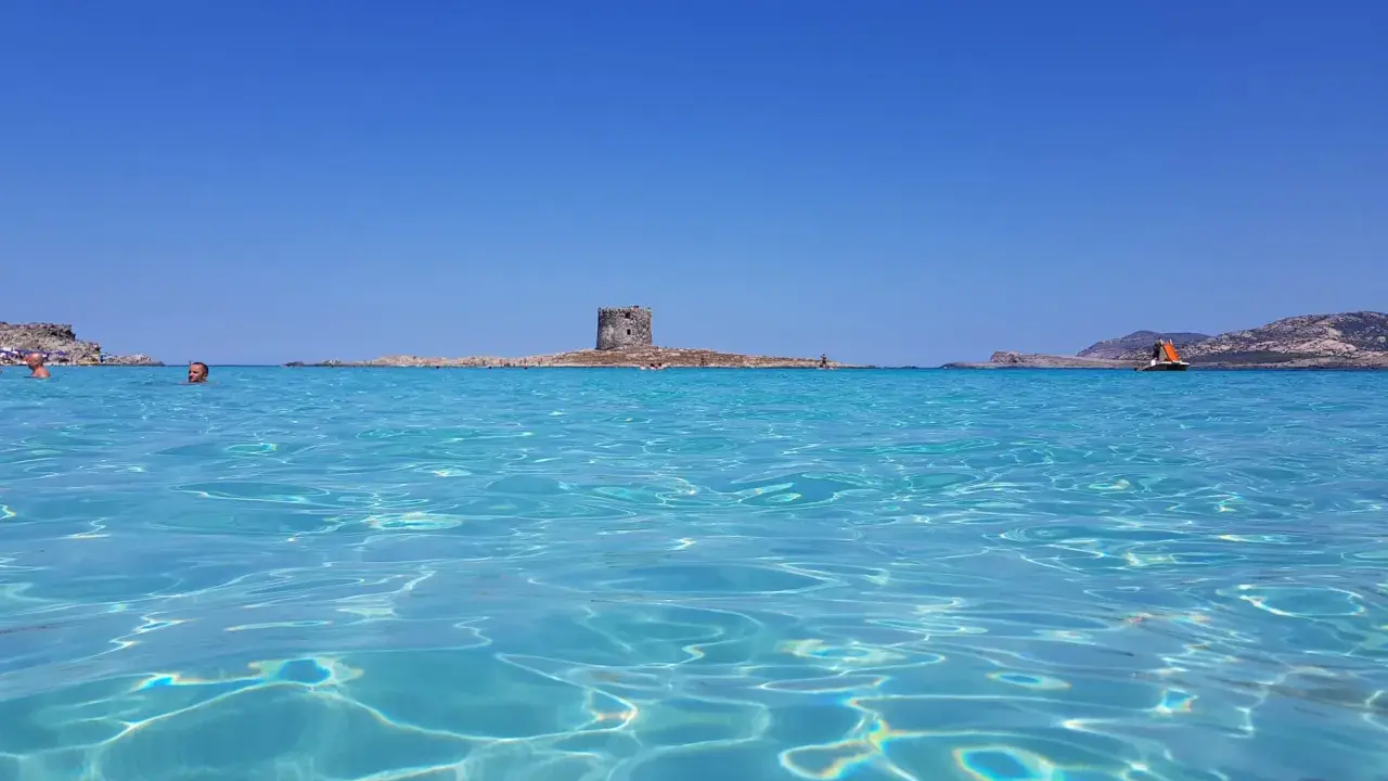 Acque cristalline del mare nord Italia con torre antica su isolotto e barca a vela arancione.