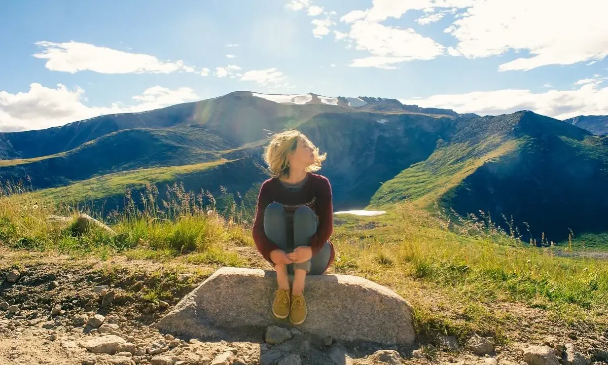 Ragazza seduta su una roccia, con i capelli mossi dal vento, ammira il panorama montano. Un momento di pace mentre camminare nella natura.