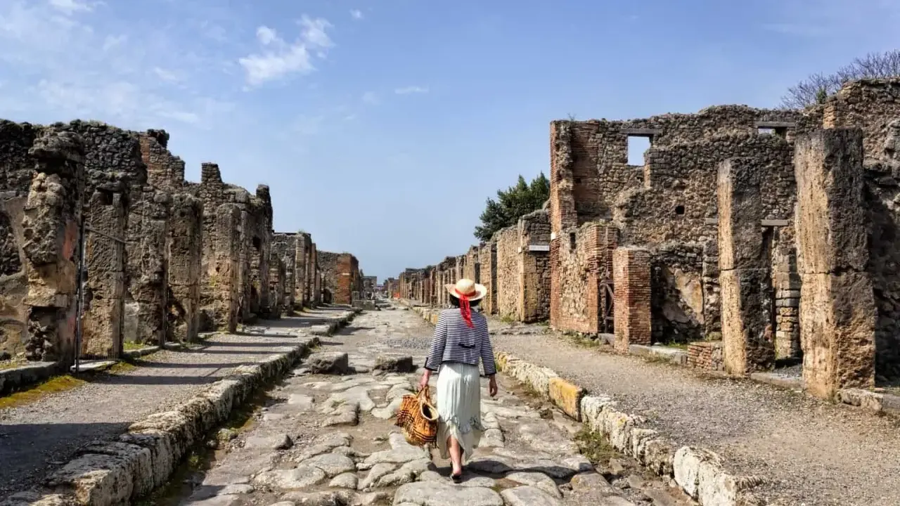 Una donna con cappello di paglia cammina lungo una strada lastricata di Pompei, tra antiche rovine. Un'esperienza unica per scoprire cosa vedere a Pompei.