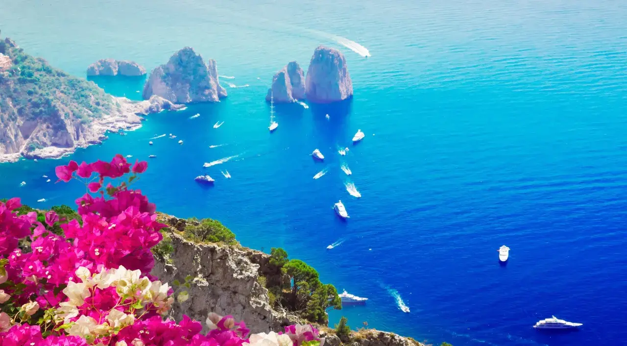 Un giorno a Capri: scogliere rocciose, mare blu intenso e barche che solcano le onde, con bouganville rosa in primo piano.