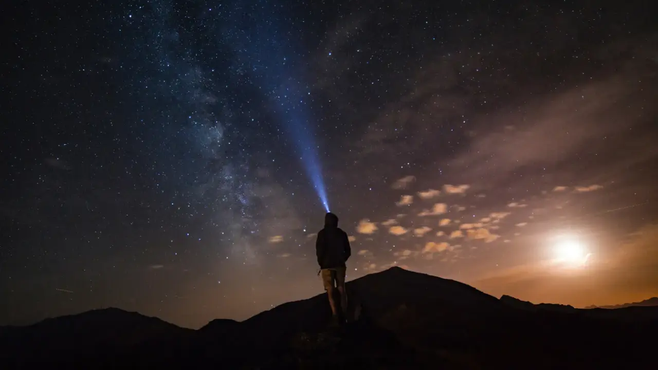 Una persona con una torcia illumina il cielo stellato su una montagna di notte. La Via Lattea è visibile.