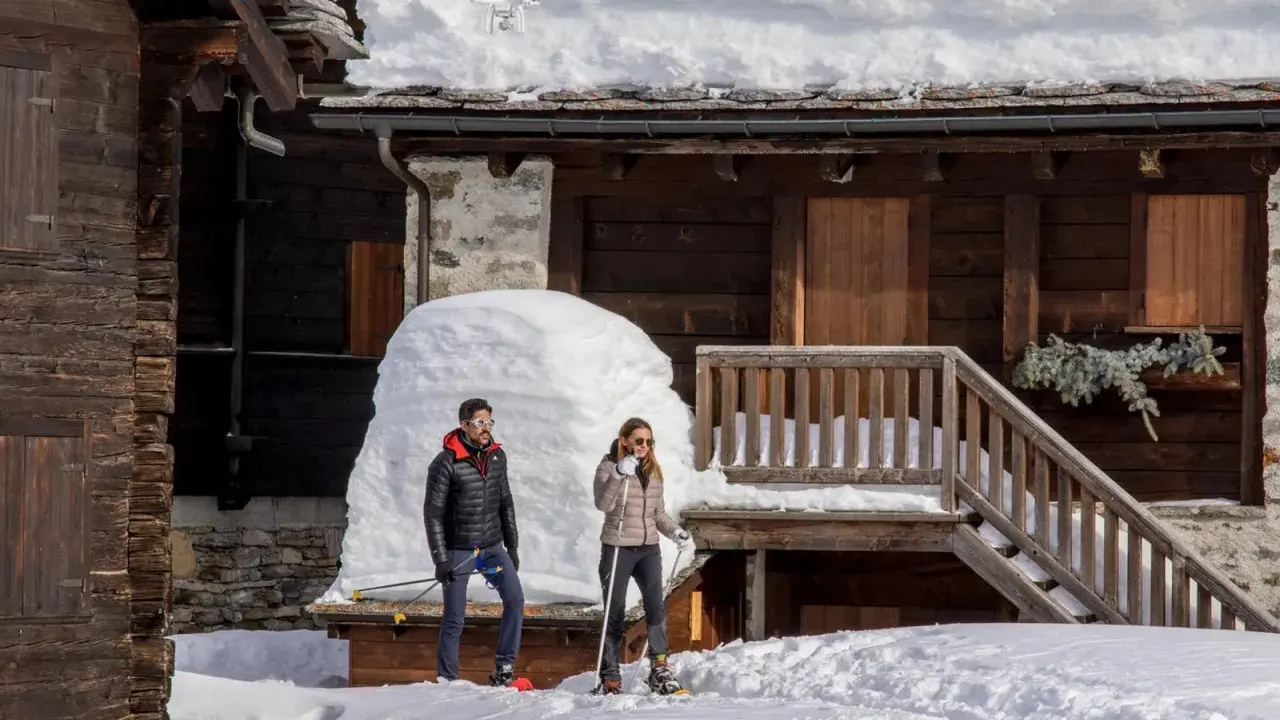 Coppia con racchette da neve a Riale Val Formazza, con un drone in volo sopra un tetto innevato.