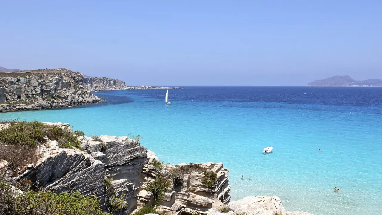 Scogliere rocciose e mare cristallino a Favignana. Un veliero solca le acque turchesi, perfetto per scoprire cosa vedere a Favignana in un giorno.