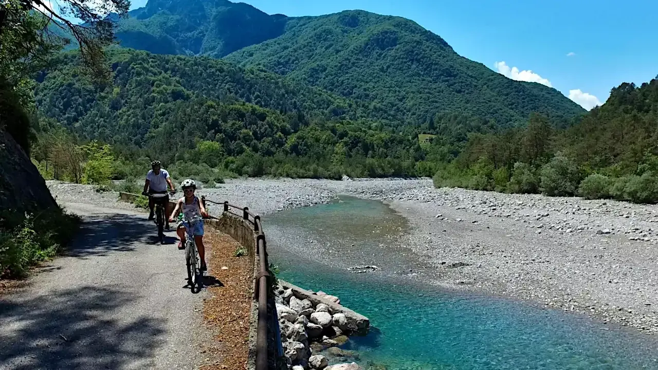 Ciclovia del Tagliamento: due ciclisti esplorano il sentiero panoramico lungo il fiume, circondati da montagne verdi e un cielo azzurro.