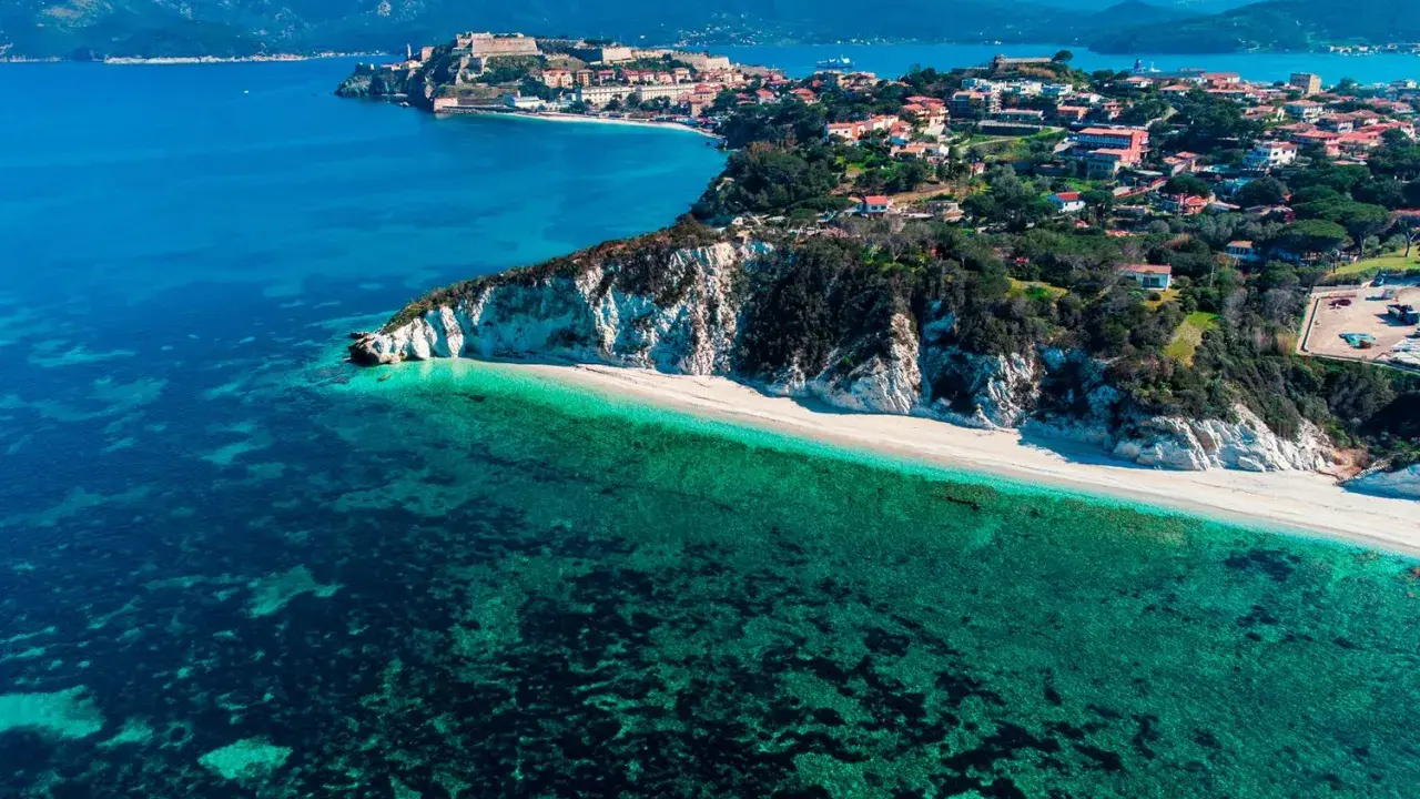 Vista aerea di una spiaggia bianca con acque cristalline e un borgo storico su una collina, parte dell'arcipelago toscano isole visitabili.