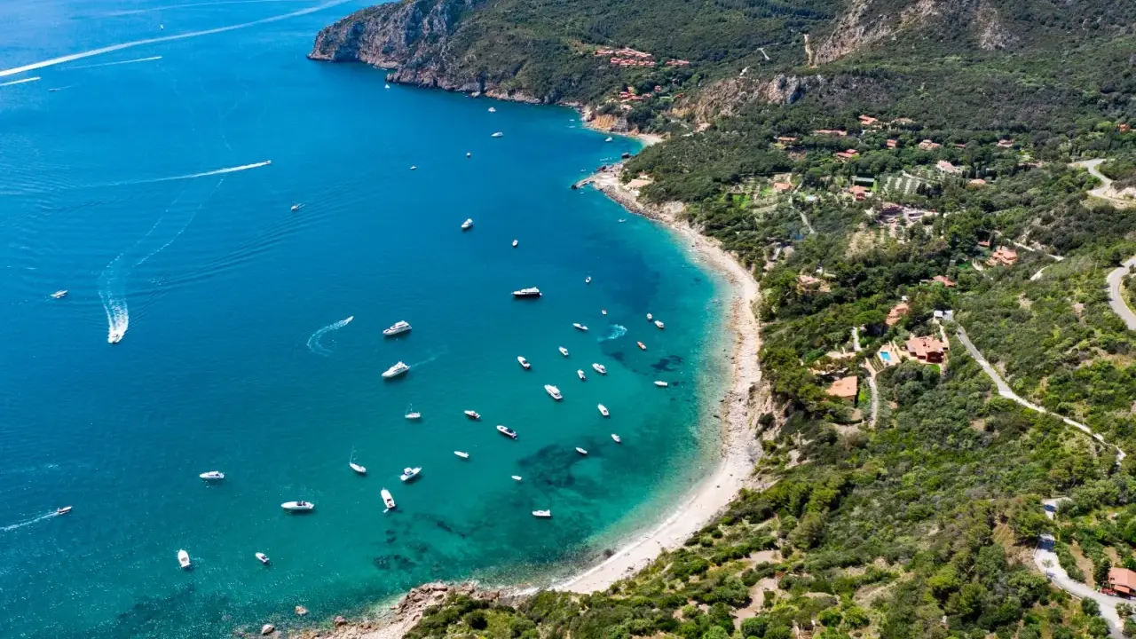 Baia con acque cristalline e numerose barche, incastonata tra colline verdi e scogliere. Un vero gioiello tra le spiagge più belle Toscana.