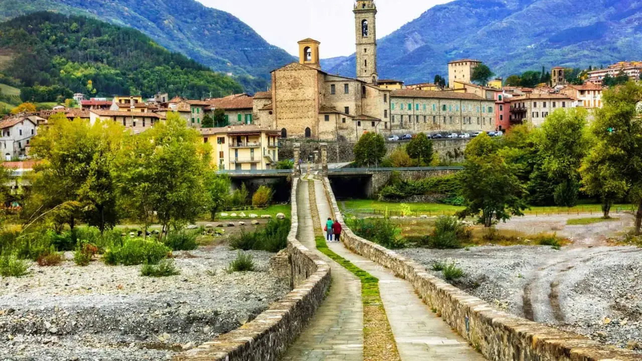 Ponte Gobbo e borghi vicino a Bobbio, con montagne sullo sfondo. Due persone passeggiano sul ponte antico.