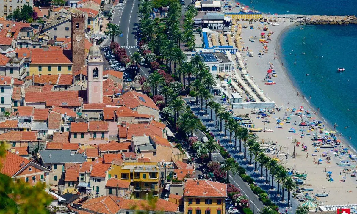 Aerial view of a coastal town with red-tiled roofs, a beach with umbrellas, and palm trees lining the road.