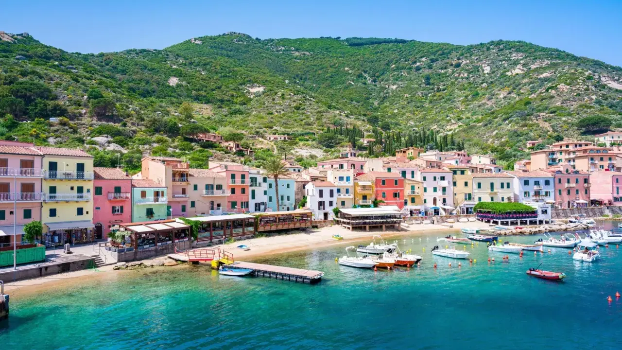 Colorate case affacciate sul mare cristallino, con barche ormeggiate e la collina verde sullo sfondo. Un'immagine che evoca il fascino del traghetto per l'Isola del Giglio.