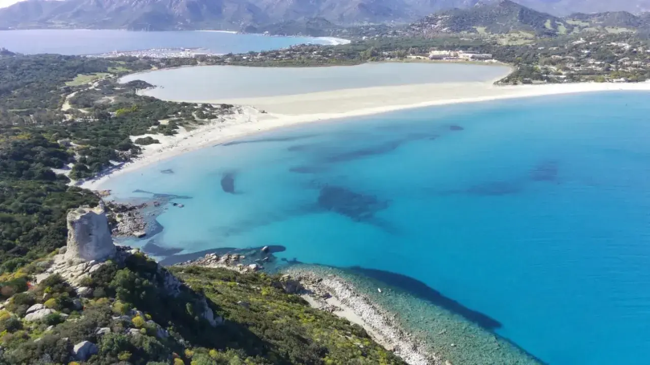 Torre costiera su promontorio verdeggiante domina una baia con acque cristalline, tra le spiagge più belle Sardegna.