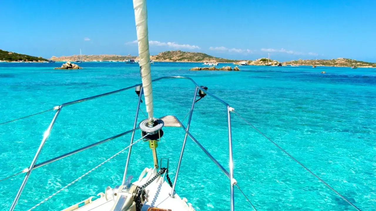 La prua di una barca a vela naviga verso La Maddalena, tra acque cristalline e isole rocciose.