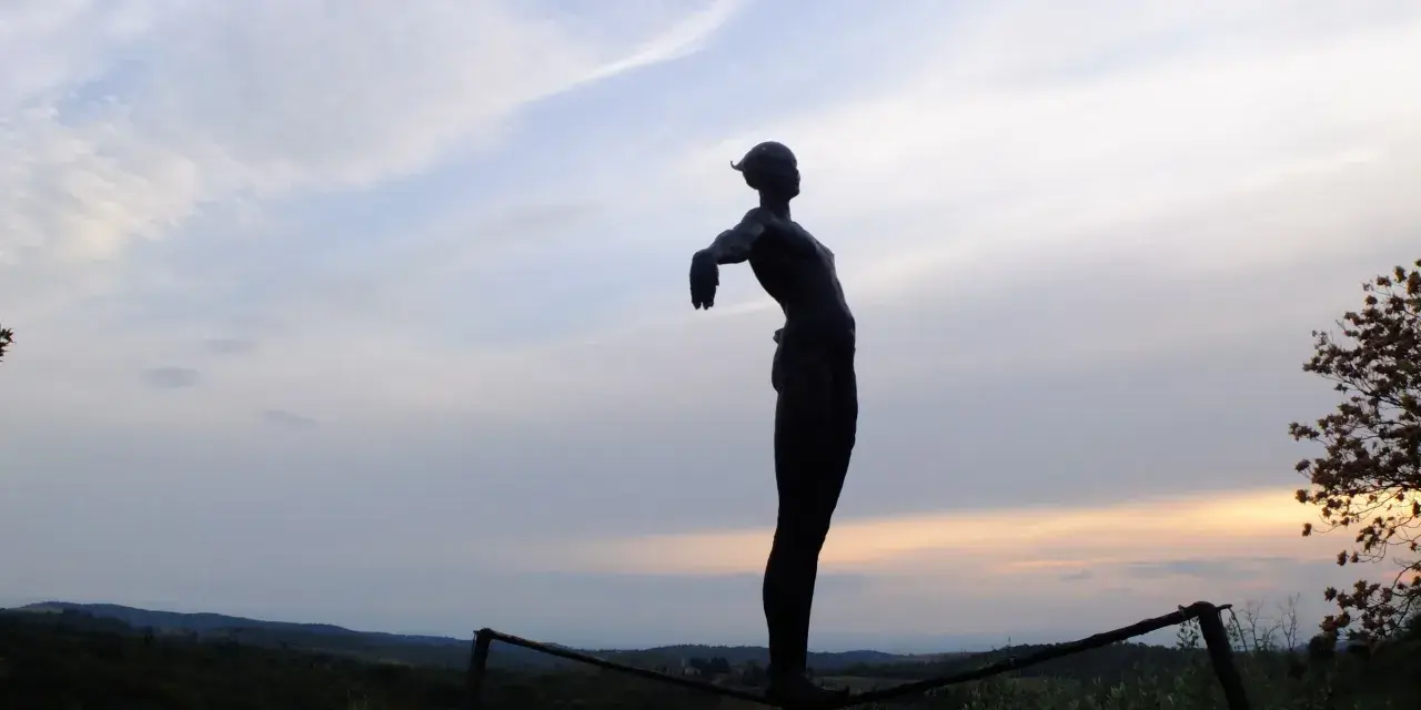 Statua silhouette con braccia aperte, su un crinale collinare, al tramonto. Un punto panoramico lungo le tappe della Via Romea Sanese.