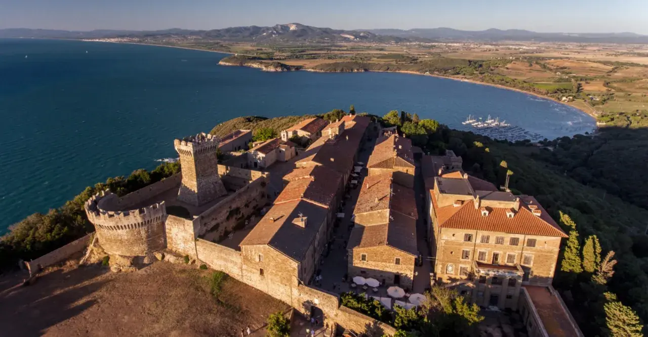 Borgo medievale fortificato domina la campagna toscana lungo il mare, con un castello imponente e case dai tetti rossi affacciate su un golfo con barche a vela.