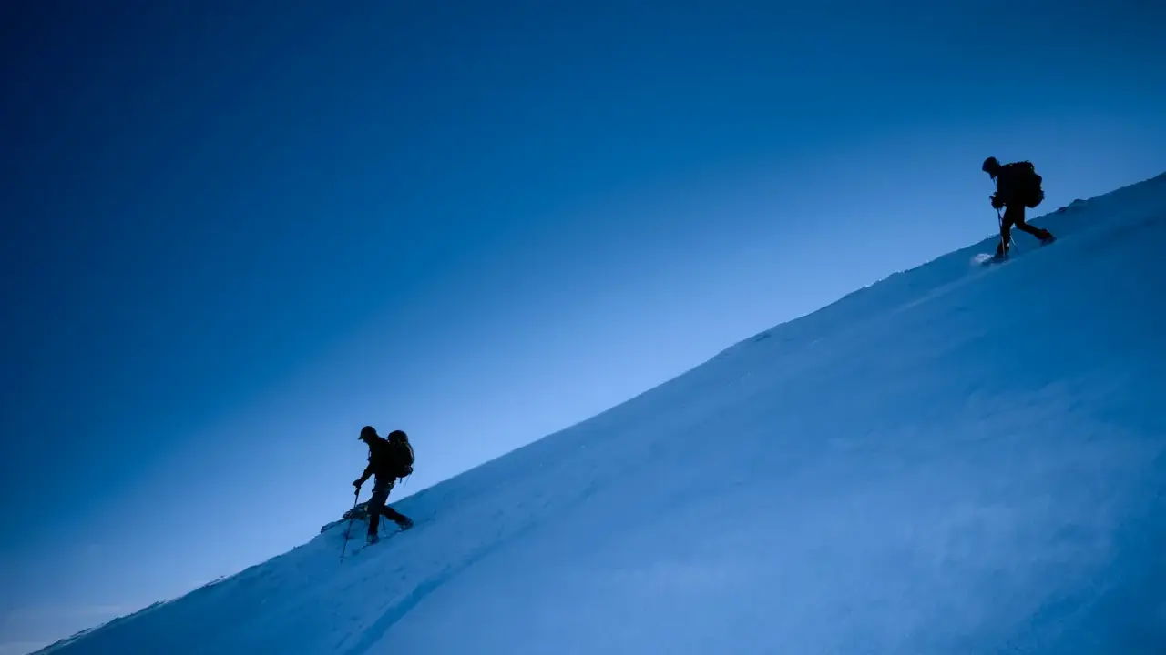 Due escursionisti con ciaspole salgono un pendio innevato sotto un cielo blu intenso. Le ciaspole sono perfette per camminare sulla neve fresca.