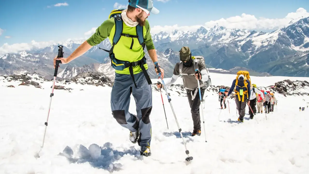 Un gruppo di escursionisti sta camminando sulla neve in montagna, con bastoncini da trekking e zaini, sotto un cielo azzurro.