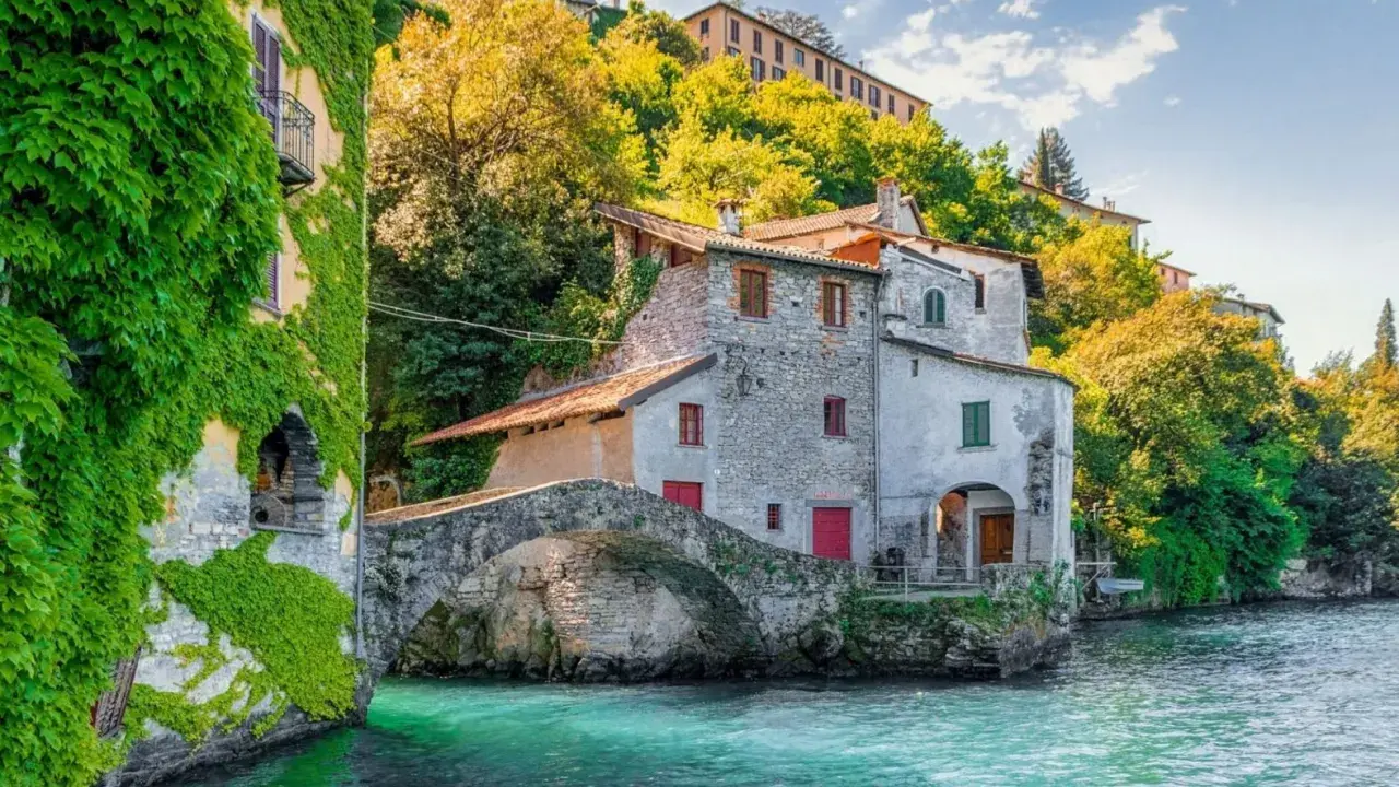 Antichi borghi medievali lombardia da visitare: un ponte di pietra collega case storiche sull'acqua cristallina, con edera rampicante e vegetazione lussureggiante.